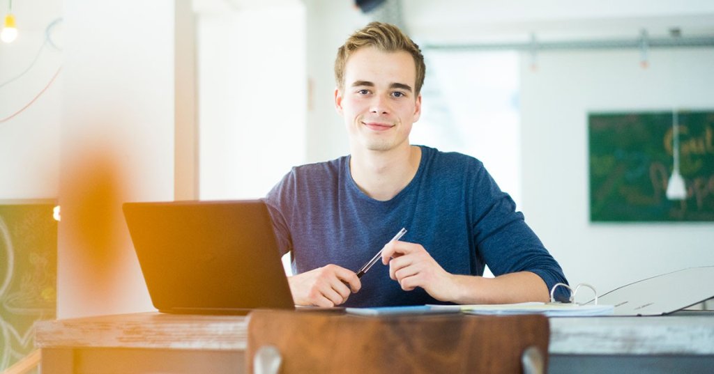 Ein junger Mann sitzt mit einem Stift in der Hand vor einem Ordner und einem Laptop und schaut grinsend in die Kamera.