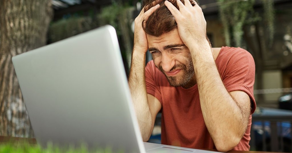 Ein Mann schlägt vor einem Laptop sitzend die Hände über dem Kopf zusammen.