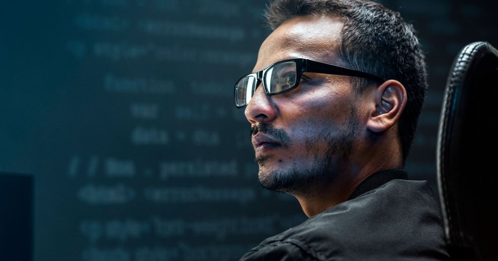 Mann mit Brille im Seitenprofil, hinter dem an der Wand ein verschwommener Code zu sehen ist.