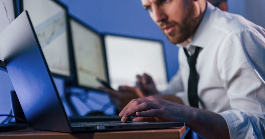 Ein Mann in weißem Hemd und scharzer Krawatte sitzt vor vielen Monitoren und schaut mit angestrengter Mimik in seinen Laptop.