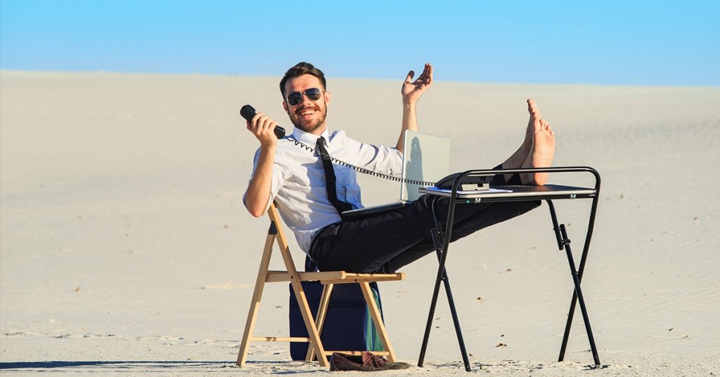 Mann mit Telefon in der Hnad und Laptop auf dem Schoß sitzt auf einem Klappstuhl in der Wüste und hat seine Füße auf eine erhöhte Ablage abgelegt.